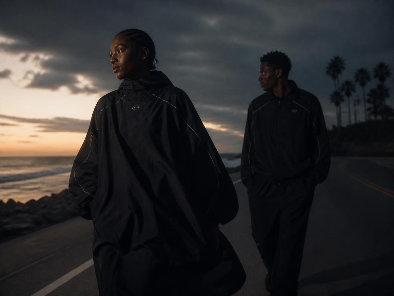Two models in dark technical outerwear on a coastal road at dusk